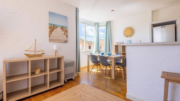 A dining area with a table and chairs, a wooden console table with a ship model, a radiator, and a window with curtains. - Comfort & Charm – 1-Min Walk to the Sea (Valras-Plage)