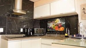 Une cuisine moderne avec des murs en marbre noir, des meubles blancs, une machine à café, un four à micro-ondes et une nature morte représentant des fruits.