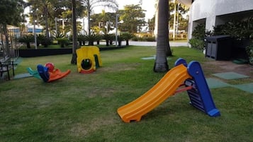 Un parque infantil con toboganes y estructuras de escalada en un área verde con palmeras y un edificio al fondo.