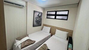 Une petite chambre à coucher, bien rangée, avec deux lits simples, un luminaire au plafond, une fenêtre et un climatiseur fixé au mur.