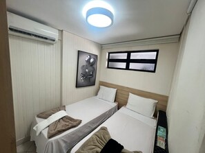 Een kleine, verzorgde slaapkamer met twee eenpersoonsbedden, een plafondlamp, een raam en een luchtverkoeler aan de muur.