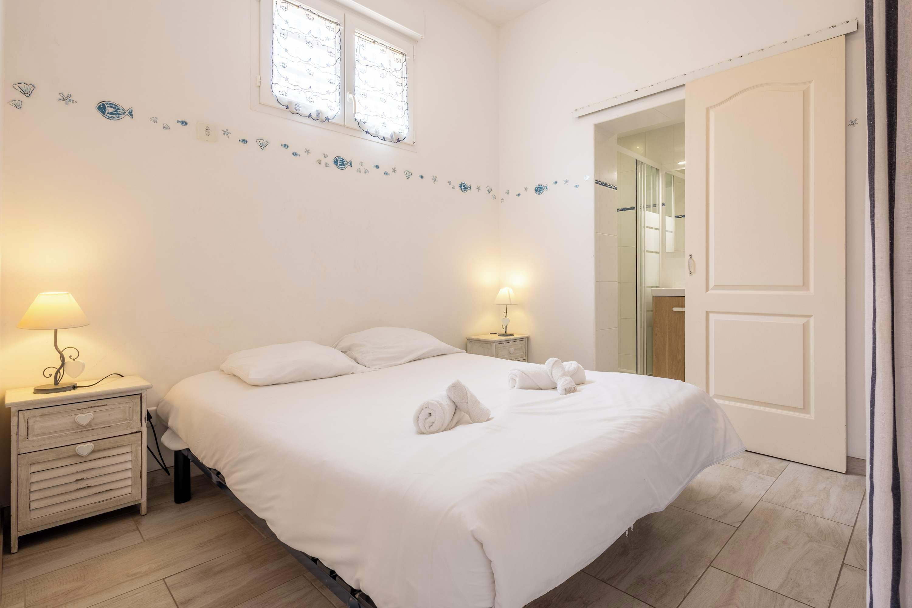 Una cama bien hecha con ropa de cama y toallas blancas, dos lámparas de noche, una mesita de noche de madera con cajones, un detalle decorativo en la pared y una puerta que da a un baño.
