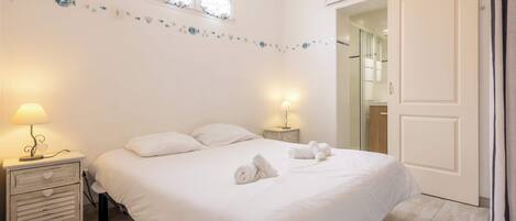 Una cama bien hecha con ropa de cama y toallas blancas, dos lámparas de noche, una mesita de noche de madera con cajones, un detalle decorativo en la pared y una puerta que da a un baño.