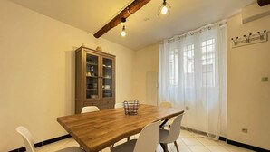 A dining area with a wooden table, white chairs, a cabinet with glass doors, and a window with curtains. - Orb Room – 1st Floor (Béziers)