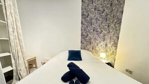 Ein aufgeräumtes Bett mit aufgerollten Handtüchern und Willkommensgeschenken, ein Nachttisch mit Lampe und eine strukturierte Wand.