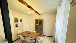Un comedor con una mesa de madera y sillas blancas, un mueble con puertas de cristal, y un techo con vigas a la vista y luces colgantes.