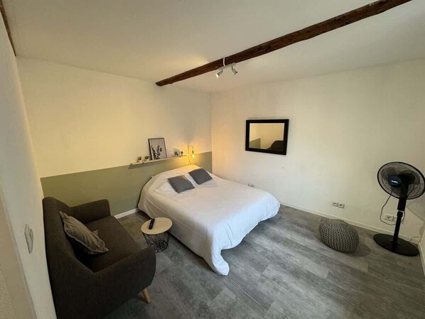 A bedroom with a bed, a sofa, a small round table, a standing fan, and a wall-mounted light fixture.