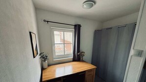 Una habitación pequeña y funcional con un escritorio de madera, una ventana que ofrece vistas a edificios, una planta en maceta y un espacio para la ducha.