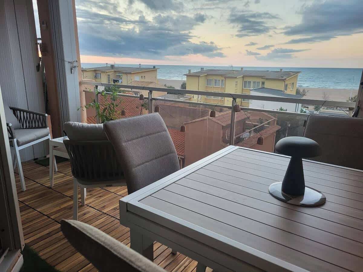 A balcony with a view of the beach and houses, a table, and two chairs.