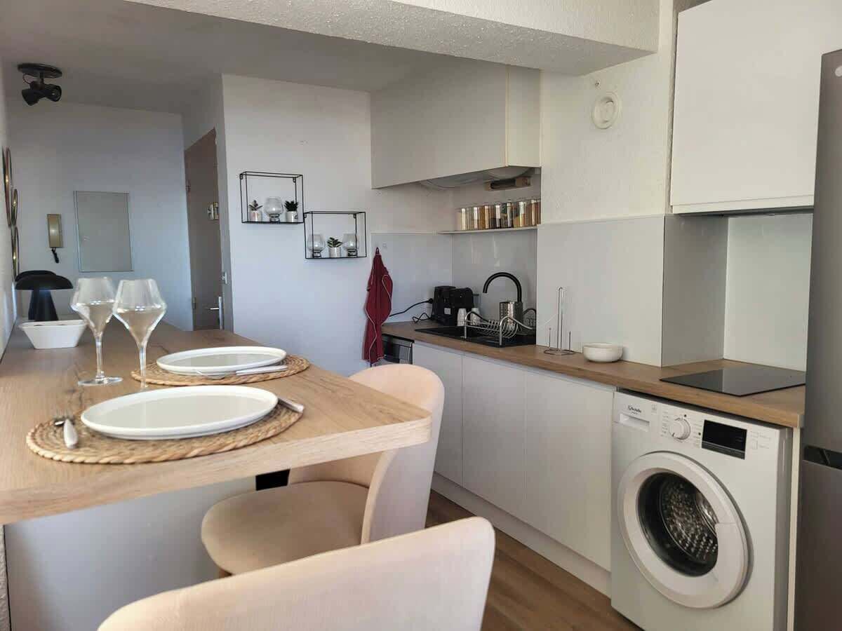 A compact kitchen with a washing machine, a dining table set with plates and glasses, and a small countertop with a kettle and jars.