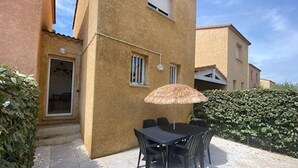 A patio with a table and chairs, a thatched umbrella, and a building with a door and windows.