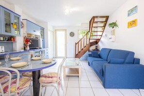 A modern living room with a blue sofa, a dining table with chairs, a kitchen area with cabinets, and a staircase.