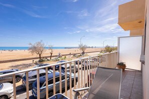 Un balcón con vista a una playa, coches aparcados y una planta en maceta.