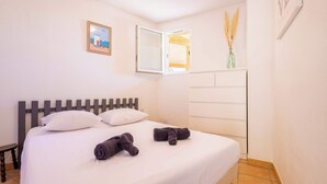 A neatly made bed with white linens and two rolled towels, a wooden headboard, a small bedside table, a window with blinds, and a wall-mounted shelf with decorative items. - T2 with Pool, Terrace & Parking (Valras-Plage)