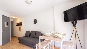 A living room with a sofa, a dining table set with plates and glasses, a mounted TV, and a wall clock. - T2 with Pool, Terrace & Parking (Valras-Plage)
