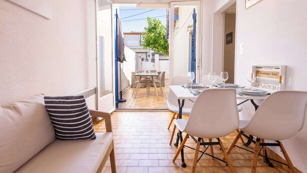 A dining area with a table set for two, a sofa with a striped pillow, and an open door leading to a courtyard.