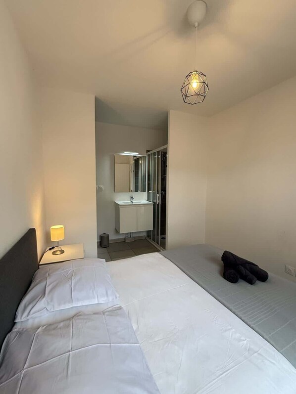 A hotel room with a bed, a bedside lamp, a small table, a mirror, and a shower area.