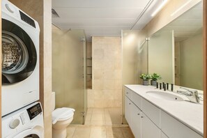 A modern bathroom with a built-in washing machine, a glass shower enclosure, a sink with a mirror, and a toilet. - Panoramic Sea View Escape by Sea N' Rent (Tel Aviv)