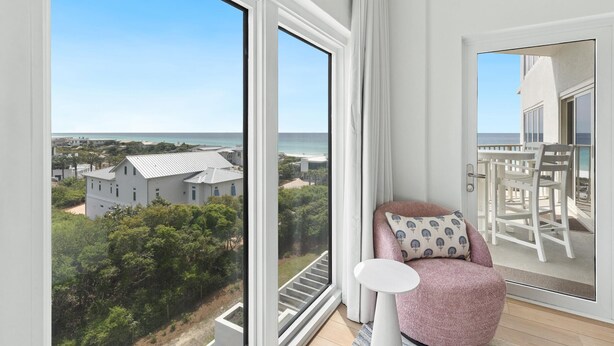 A balcony with a view of the sea and a building, a pink chair, and a white table.