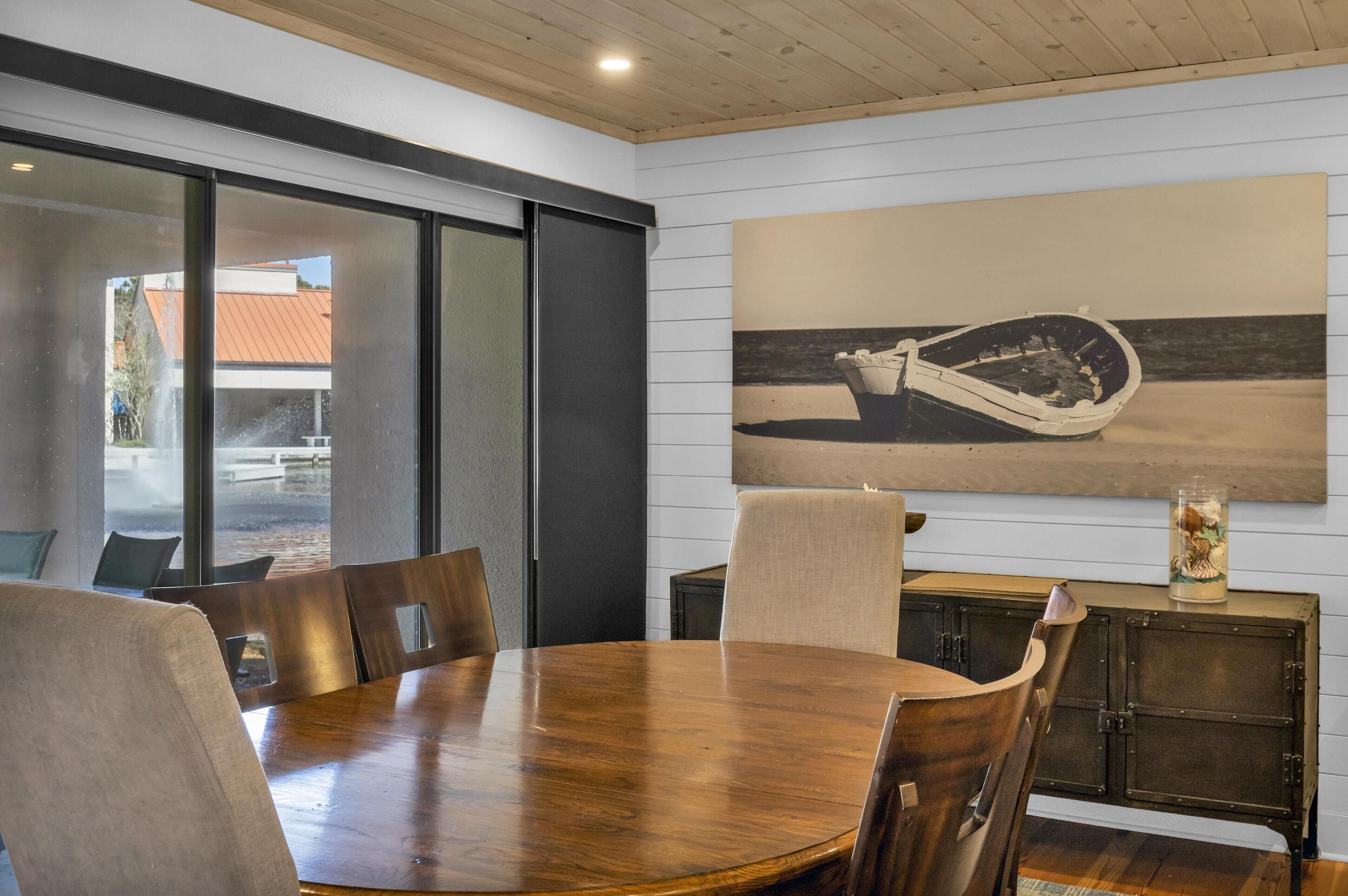 Un comedor con una mesa y sillas de madera, un ventanal grande y una obra de arte en la pared que muestra un barco.