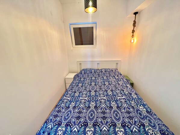 Una habitación estrecha con una cama, un ventanal pequeño y una lámpara colgante.