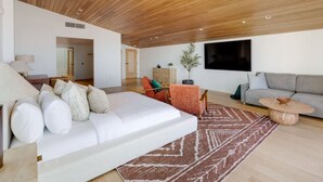 Een moderne woonkamer met een witte bank, een grijze bank en een rode fauteuil. Er is een groot raam, een houten plafond en een flatscreen televisie aan de muur bevestigd.