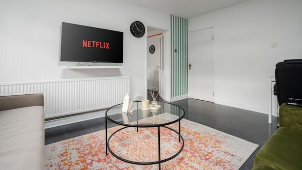 Een moderne woonkamer met een flatscreen televisie aan de muur, een ronde glazen salontafel, een bank en een groene fauteuil.