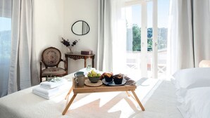A bedroom with a bed, a bedside table with a tray of food, a chair, and a view of the outdoors.