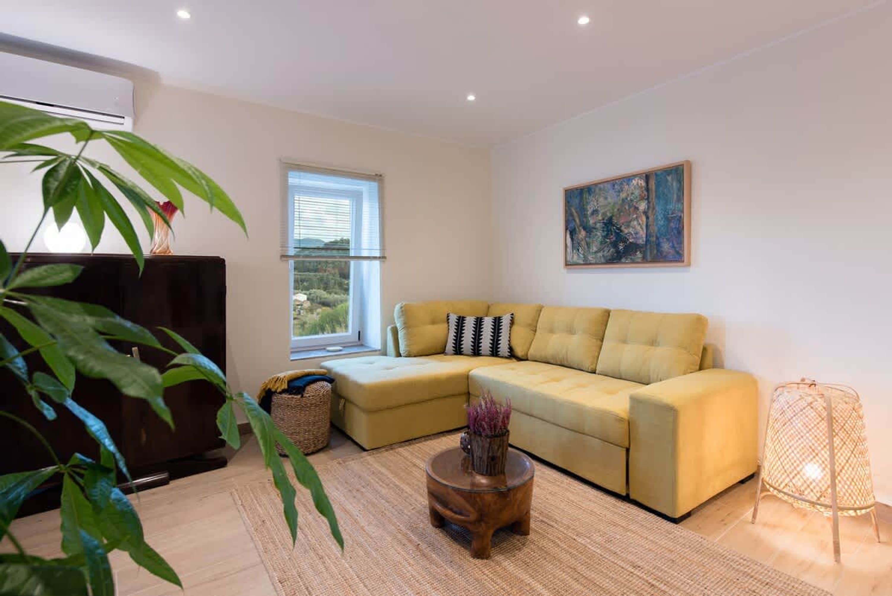 A living room with a yellow sectional sofa, a wooden coffee table, a large window with blinds, a painting on the wall, and a potted plant.