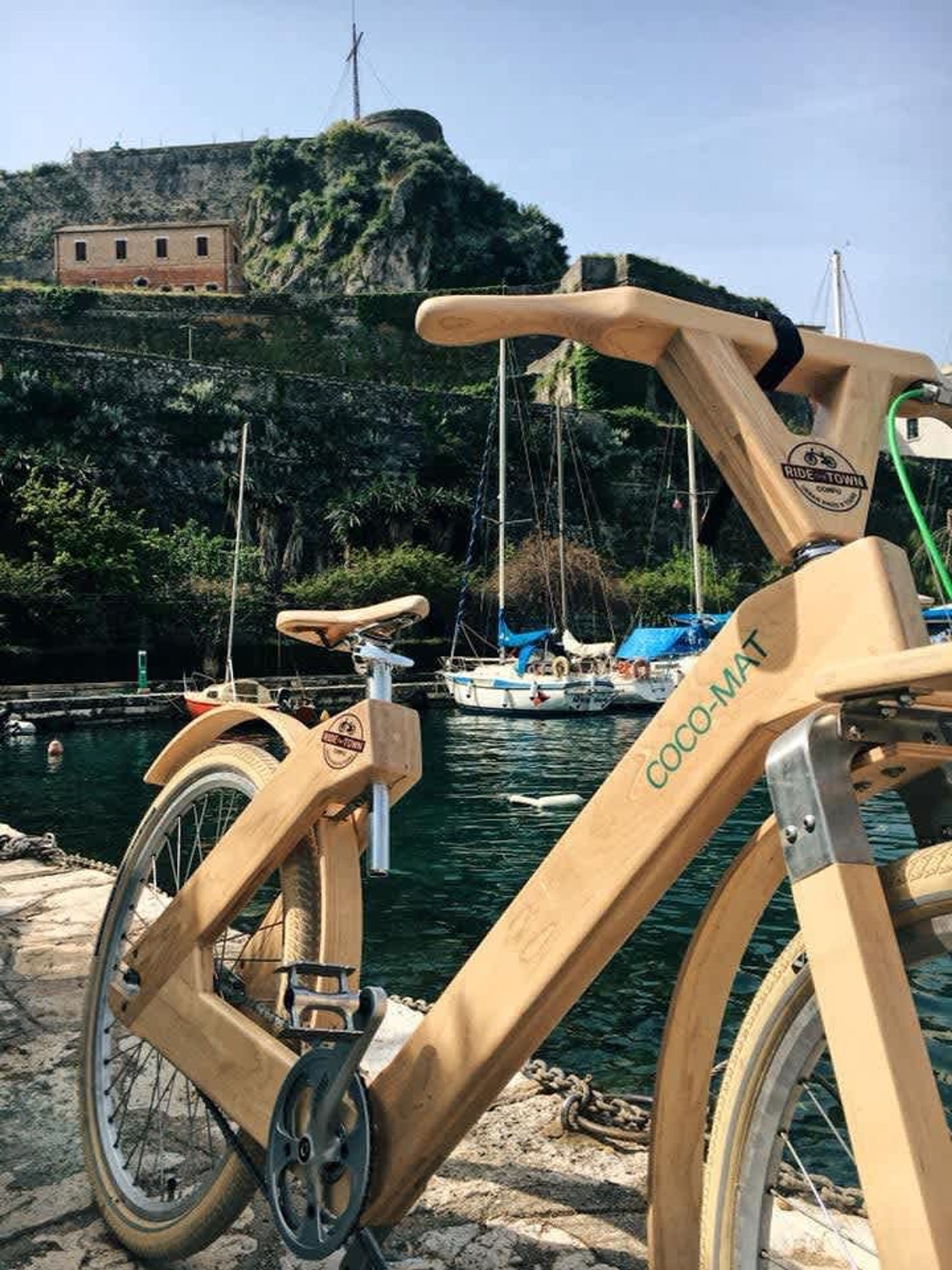 A wooden bicycle with "COCO-MAT" branding, parked by a waterfront with boats and a rocky hill in the background.