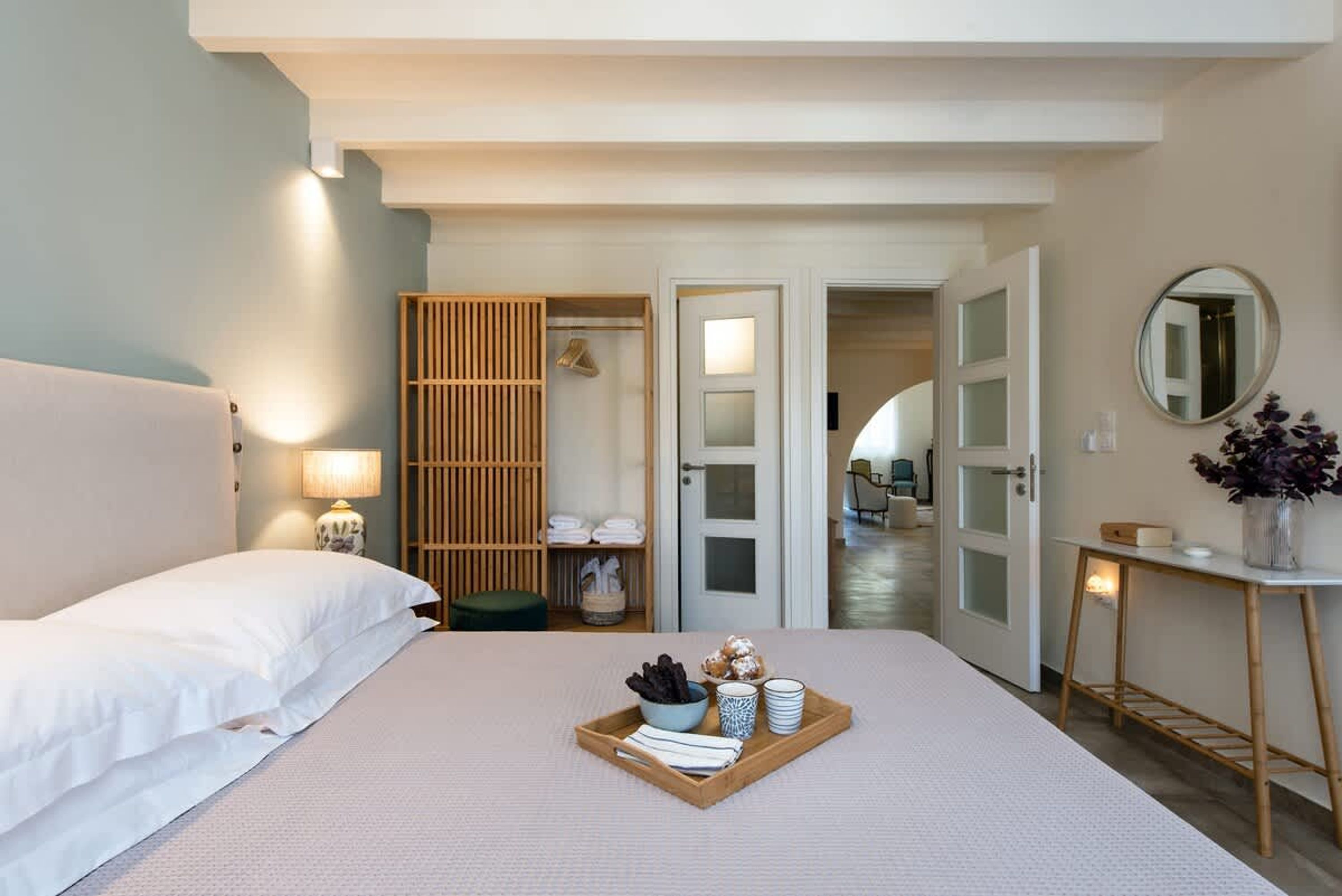 Een hotelkamer met een bed, een nachtkastje met een lamp, een dienblad met toiletartikelen, een spiegel en uitzicht op de gang.