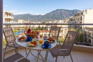 Balcón con mesa dispuesta para el desayuno, incluyendo cruasanes, fruta y jugo, con vistas a la ciudad y montañas.