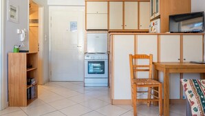 A compact kitchen with a refrigerator, oven, and wooden cabinets. There is a chair and a small table with a TV. - Comfy Beach Flat 10min to the City Center (Kalamata)