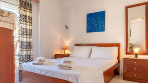A neatly made bed with white linens and towels, a bedside lamp, a wooden nightstand, a wall-mounted painting, and a mirror. - Comfy Beach Flat 10min to the City Center (Kalamata)
