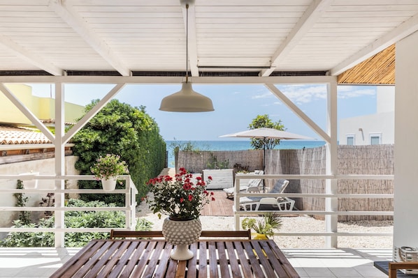A covered patio with a wooden table, a potted plant, and a view of the sea.