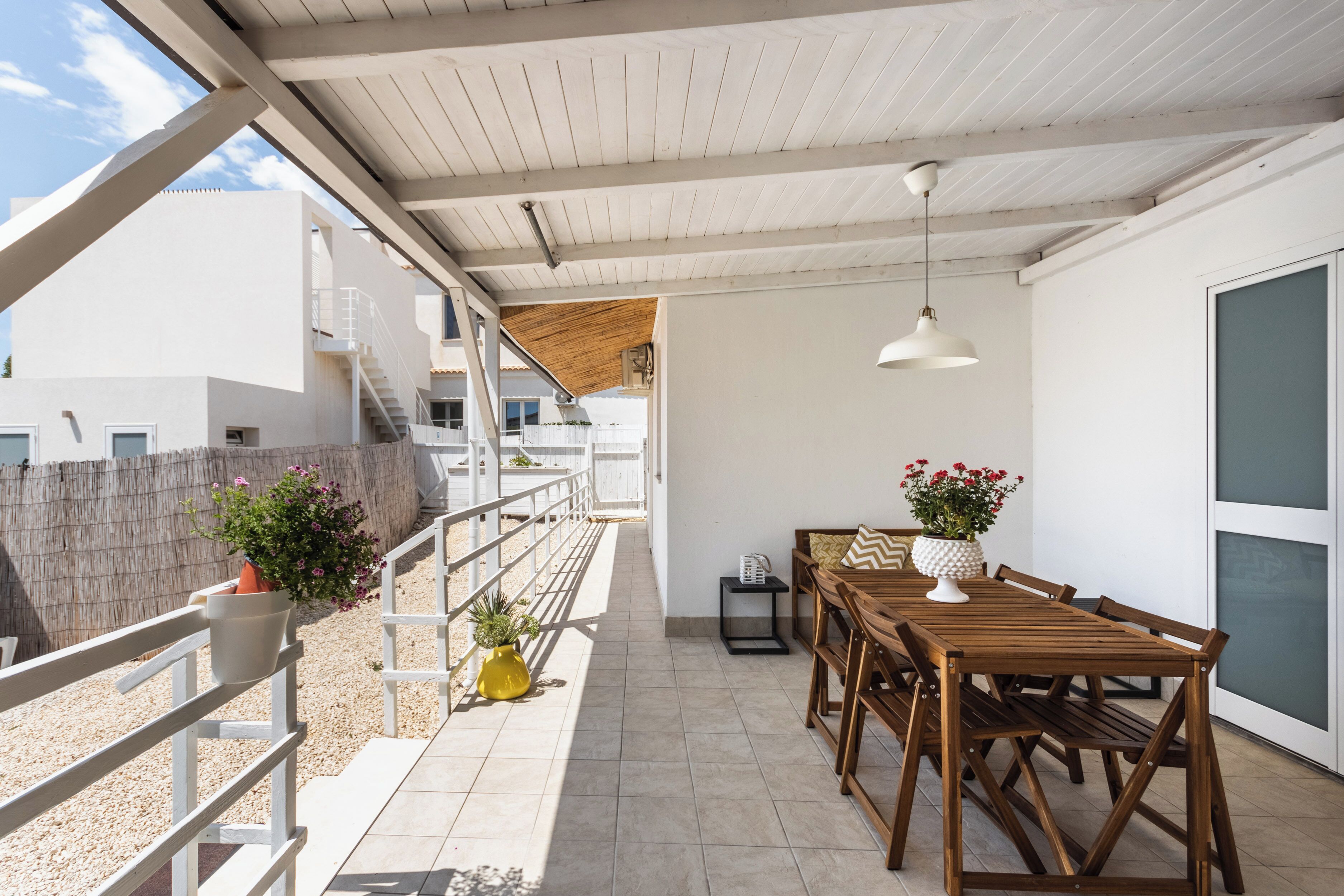 Un espacio al aire libre cubierto con una mesa y sillas de madera, plantas en macetas y una escalera que sube a un nivel superior.
