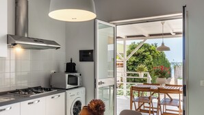 Una cocina moderna con estufa a gas, microondas y lavadora, un comedor con muebles de madera, y vistas a zonas verdes y un edificio a través de un ventanal.