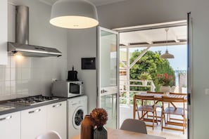 A modern kitchen with a gas stove, microwave, and washing machine, a dining area with wooden furniture, and a view of greenery and a building through a glass door.