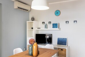 Un soggiorno moderno con un tavolo in legno, due vasi, una televisione a schermo piatto, un lampadario bianco a sospensione, una sveglia a parete e una mensola con oggetti decorativi.