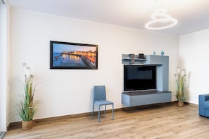 Un soggiorno moderno con una televisione a schermo piatto, un quadro incorniciato, una poltrona blu e due piante in vaso.