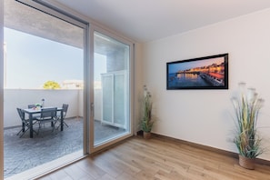 Una stanza con pavimento in legno, una porta scorrevole in vetro che dà su una zona esterna con tavolo e sedie, un quadro appeso al muro e due piante in vaso.