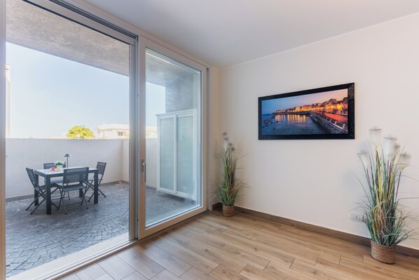Una habitación con piso de madera, una puerta corrediza de vidrio que da a un espacio exterior con una mesa y sillas, un cuadro en la pared y dos plantas en macetas.
