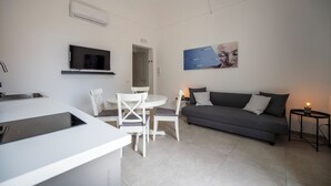 A modern living room with a grey sofa, white dining table, and chairs, a flat-screen TV mounted on the wall, and a small side table with a candle. - Demetra - One Bedroom Apartment, Sleeps 4 (Noto)