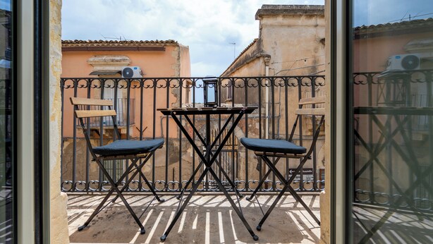 A balcony with a table and two chairs, a wrought iron fence, and buildings in the background. - Demetra - One Bedroom Apartment, Sleeps 4 (Noto)