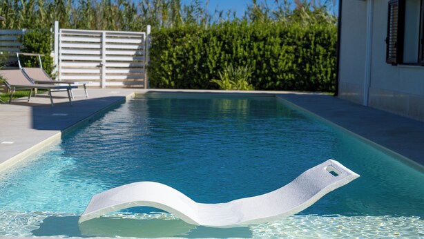 Ein Schwimmbecken mit einem weißen, schwimmenden Objekt, einem weißen Zaun und einem Gebäude mit einem Fenster.
