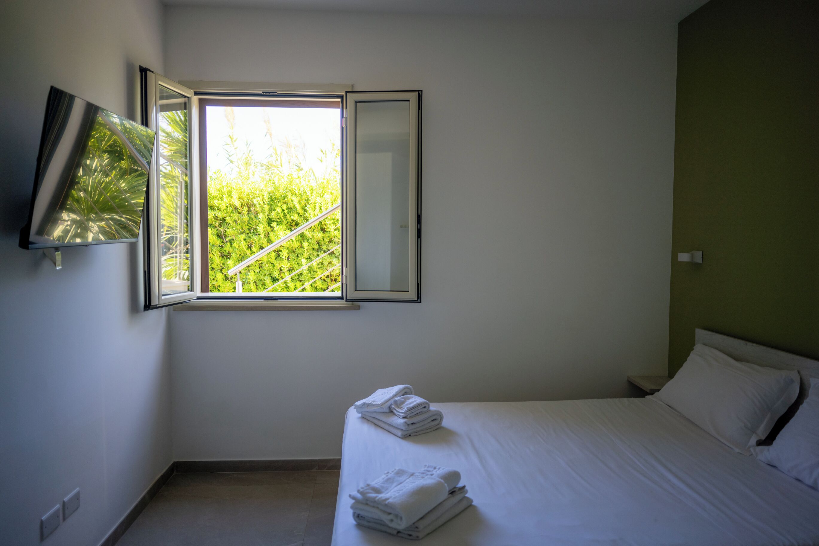 A bedroom with a bed, a window with a view of greenery, a flat-screen TV mounted on the wall, and a bedside table.