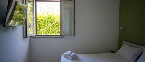 A bedroom with a bed, a window with a view of greenery, a flat-screen TV mounted on the wall, and a bedside table.
