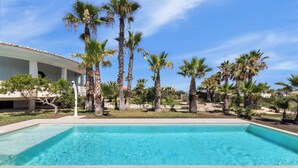 A swimming pool with clear blue water, surrounded by palm trees and a building with a tiled roof.