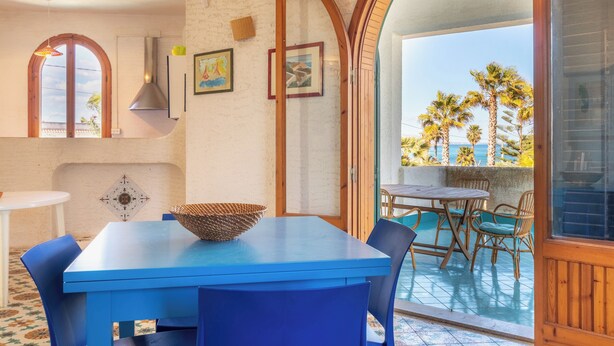 A dining area with a blue table and chairs, a wicker bowl, and a view of palm trees through an arched doorway.