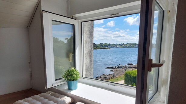 Ein Zimmer mit großem Fenster mit Blick auf ein Gewässer und eine felsige Küste. Zwei gepolsterte Bänke befinden sich im Raum.